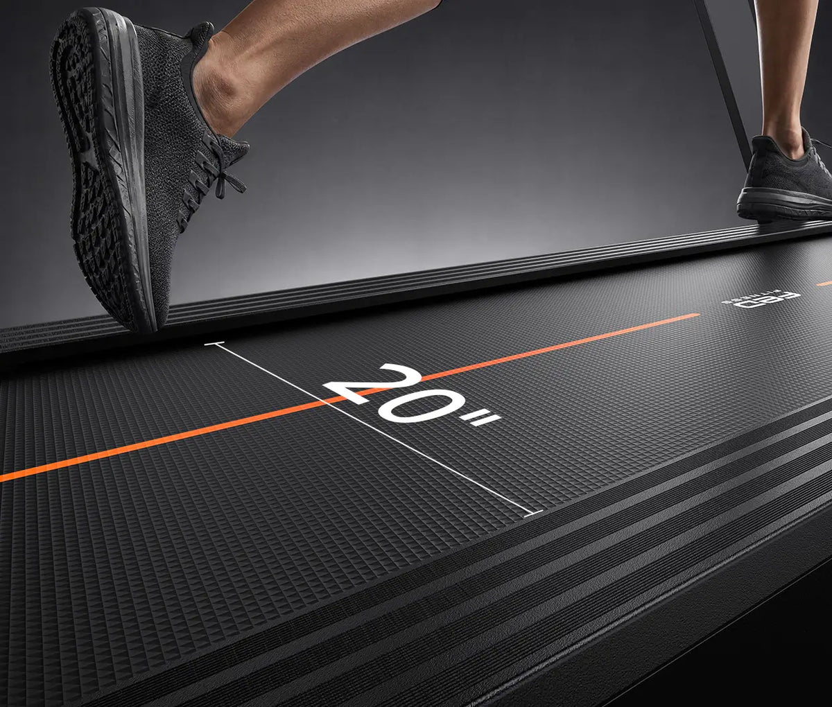 Close-up of a treadmill with a person's legs and shoes visible, showing the treadmill belt and a white measurement graphic indicating 20 inches.