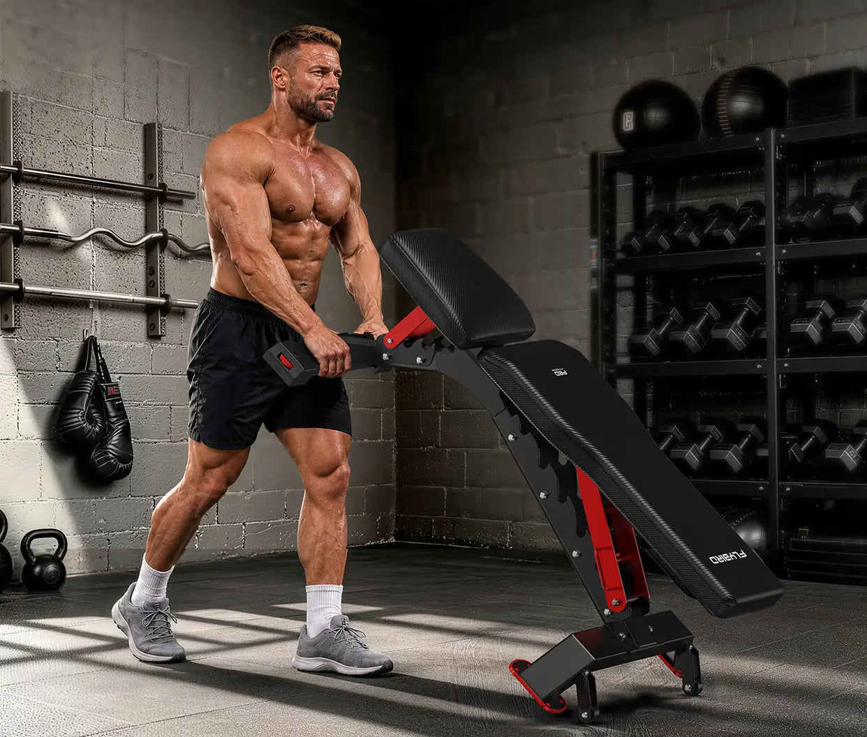 A muscular man in a gym setting pushes an adjustable black and red weight bench. The background features a gray brick wall, hanging punching bags, and a rack of dumbbells.
